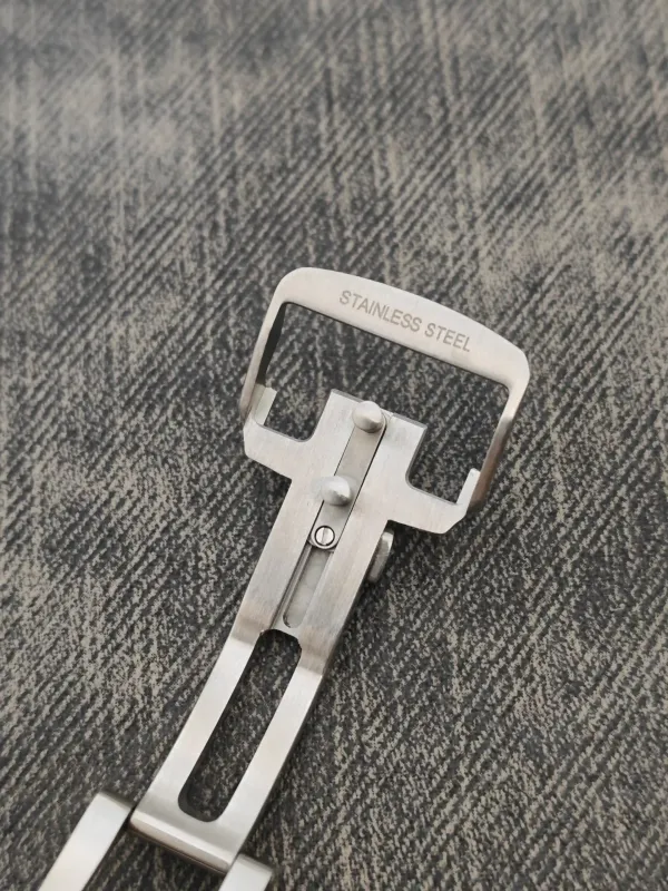 Close-up of a stainless steel buckle/clasp resting on denim, with 'STAINLESS STEEL' engraved on the top bar.