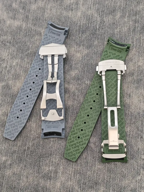 Two unfolded watch straps with stainless steel deployant buckles, one blue-gray and one green, displayed on denim fabric.
