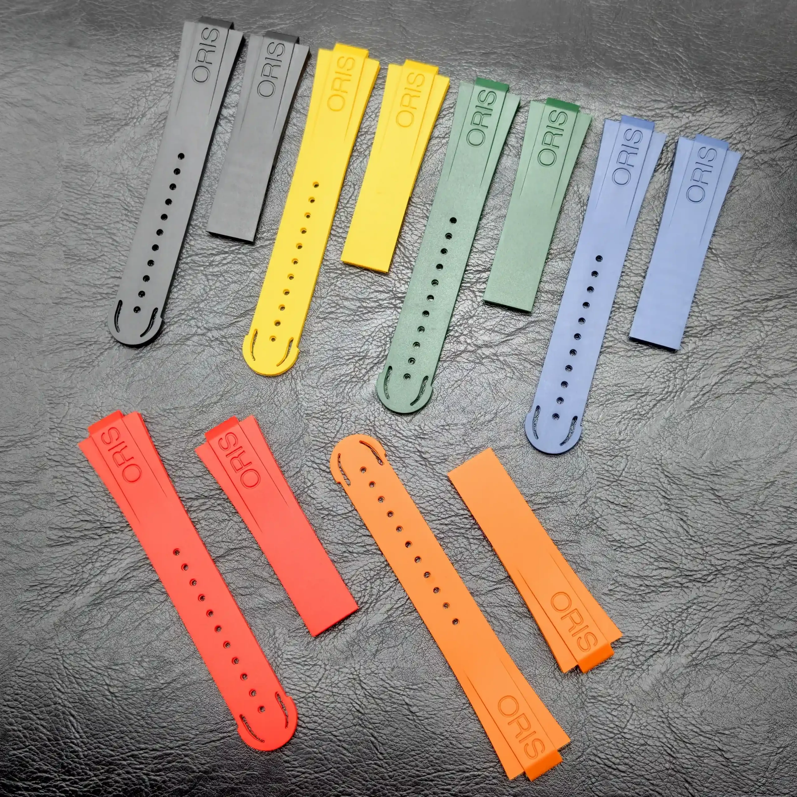 Diagonally arranged silicone watch straps in gray, yellow, green, blue, and orange with ORIS branding on each piece.