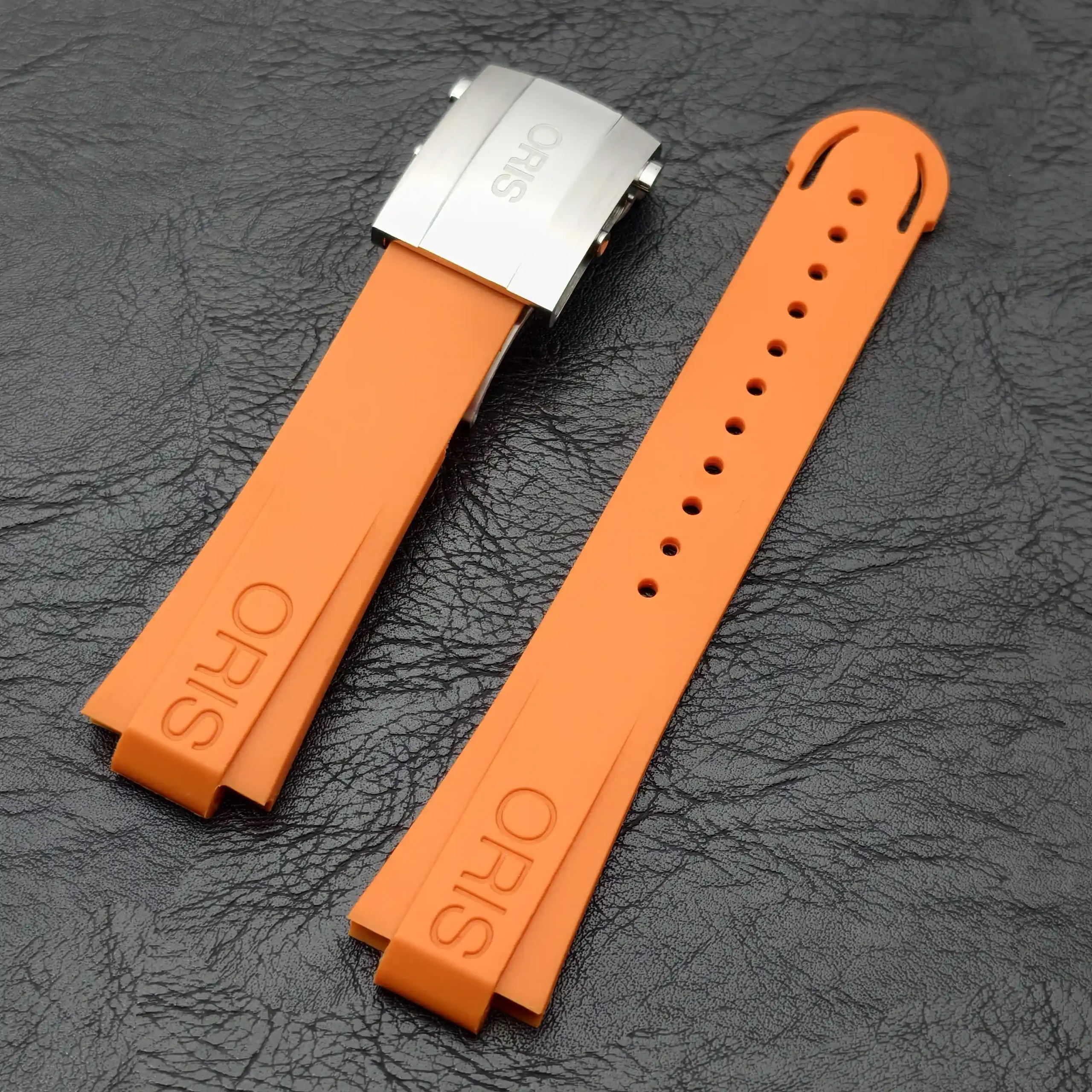 Two orange silicone watch straps with a silver metal clasp on a textured dark surface