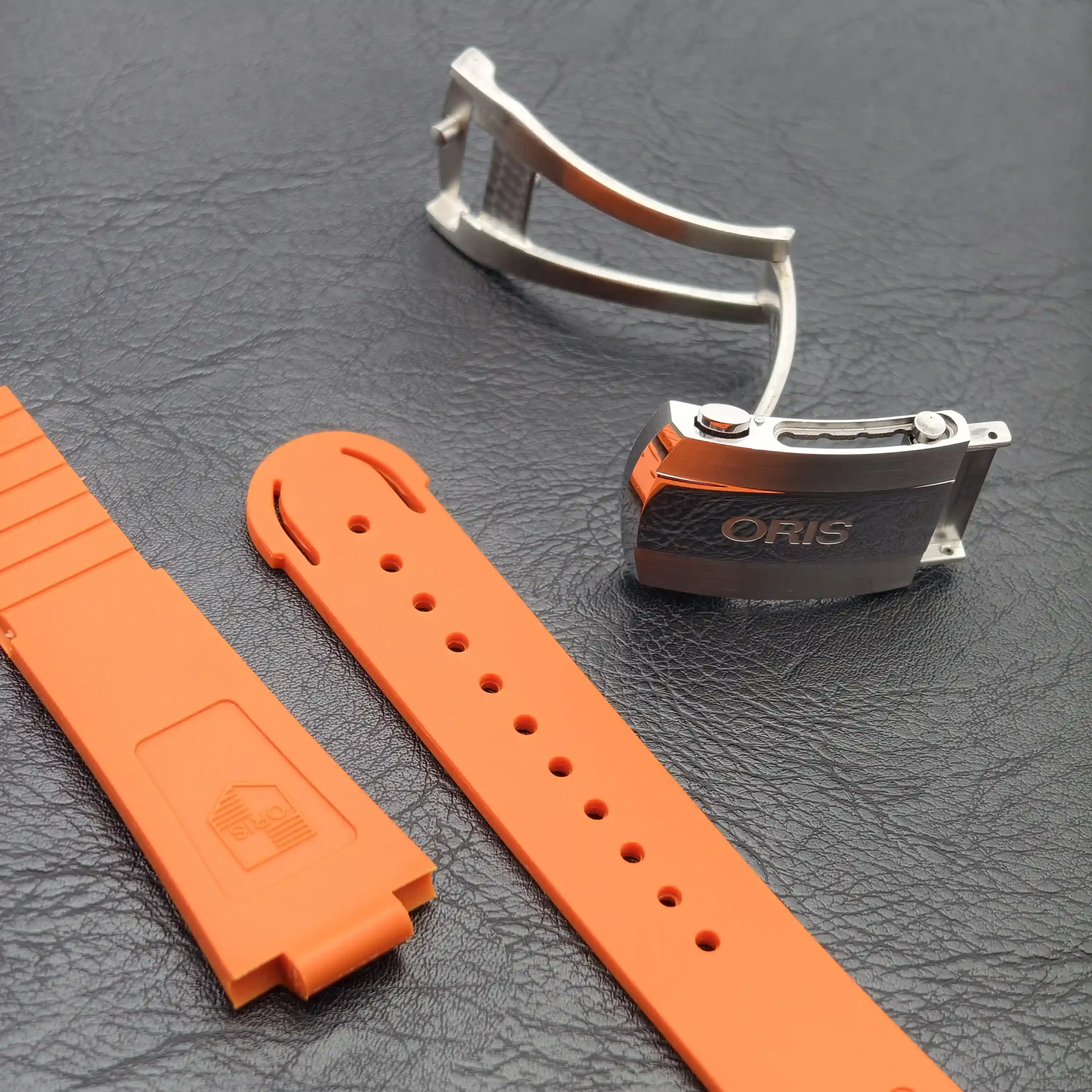 Close-up of orange silicone watch bands with holes and a silver buckle on a dark textured surface that resembles leather gores the background.