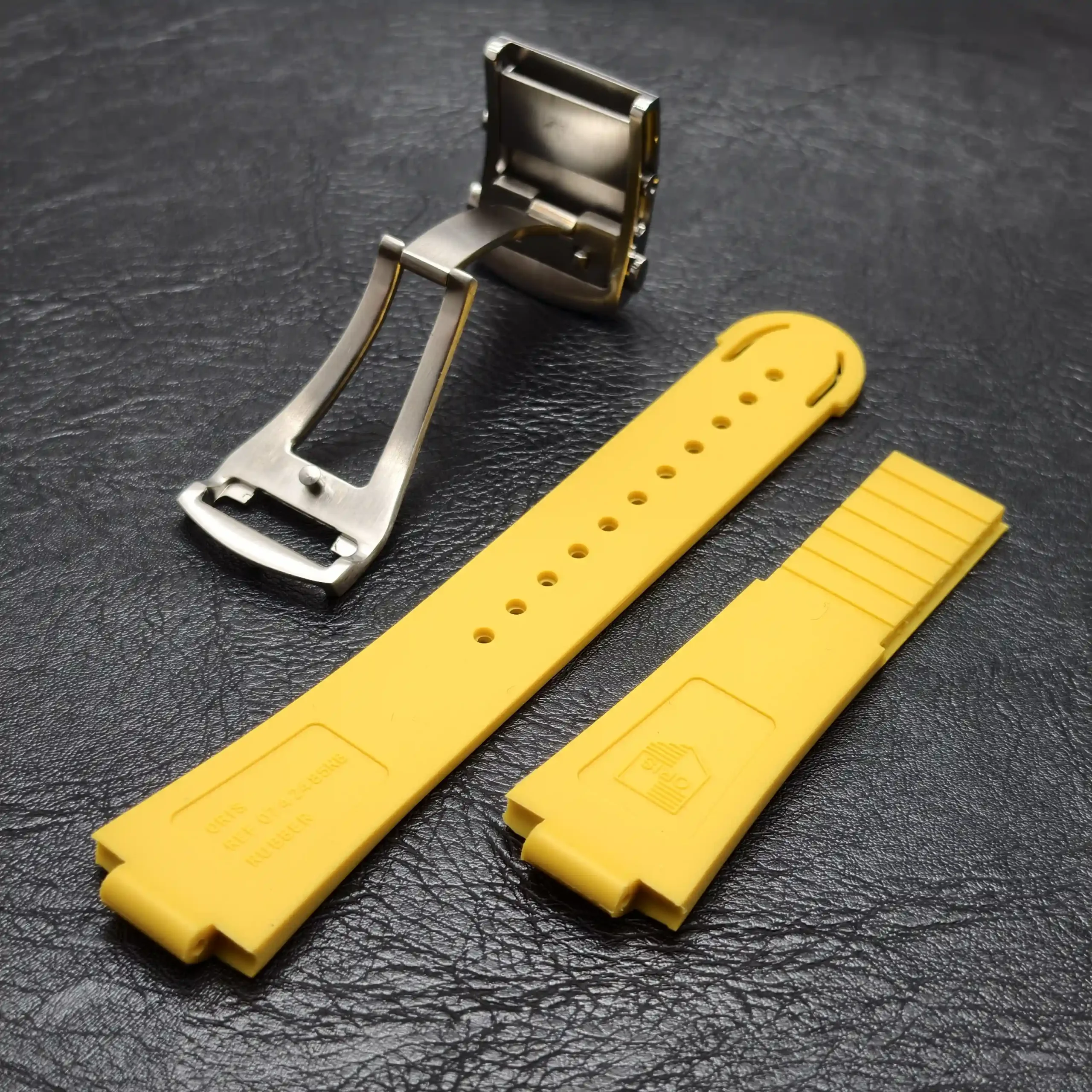 Stainless steel deployment clasp with a yellow watch strap laid out beside it on a dark textured surface
